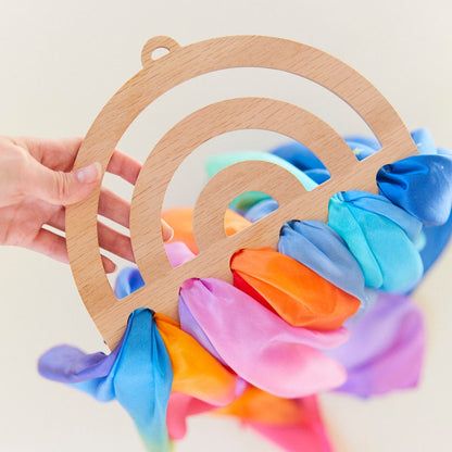 A hand holds a wooden Rainbow Playsilk Display, with colorful silk scarves threaded through it—perfect for brightening up any playroom.